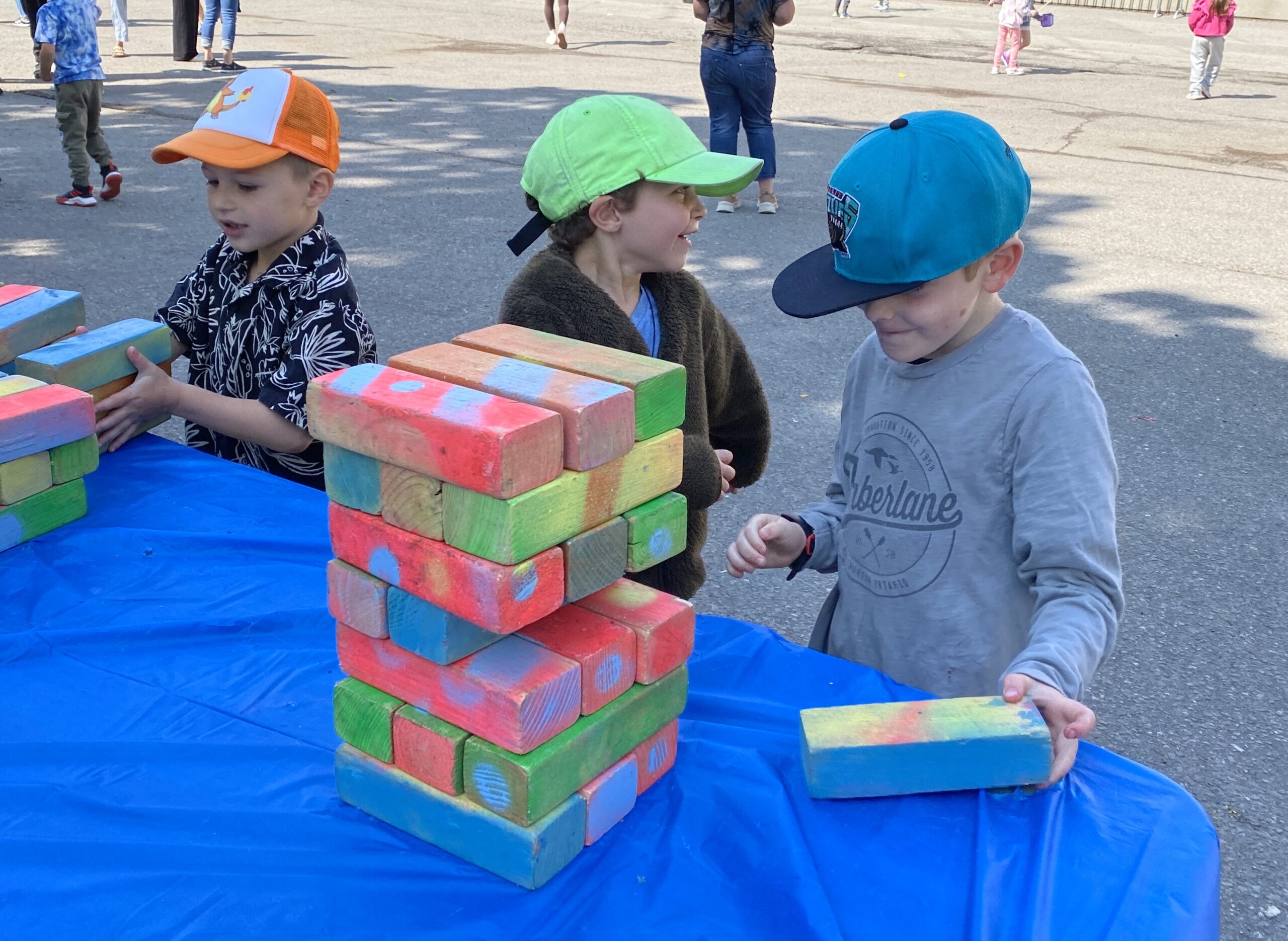 Three children playing outdoors on a tarmac with a Jenga-style game set up on a table. The wooden blocks are spray painted and one boy is taking his turn.