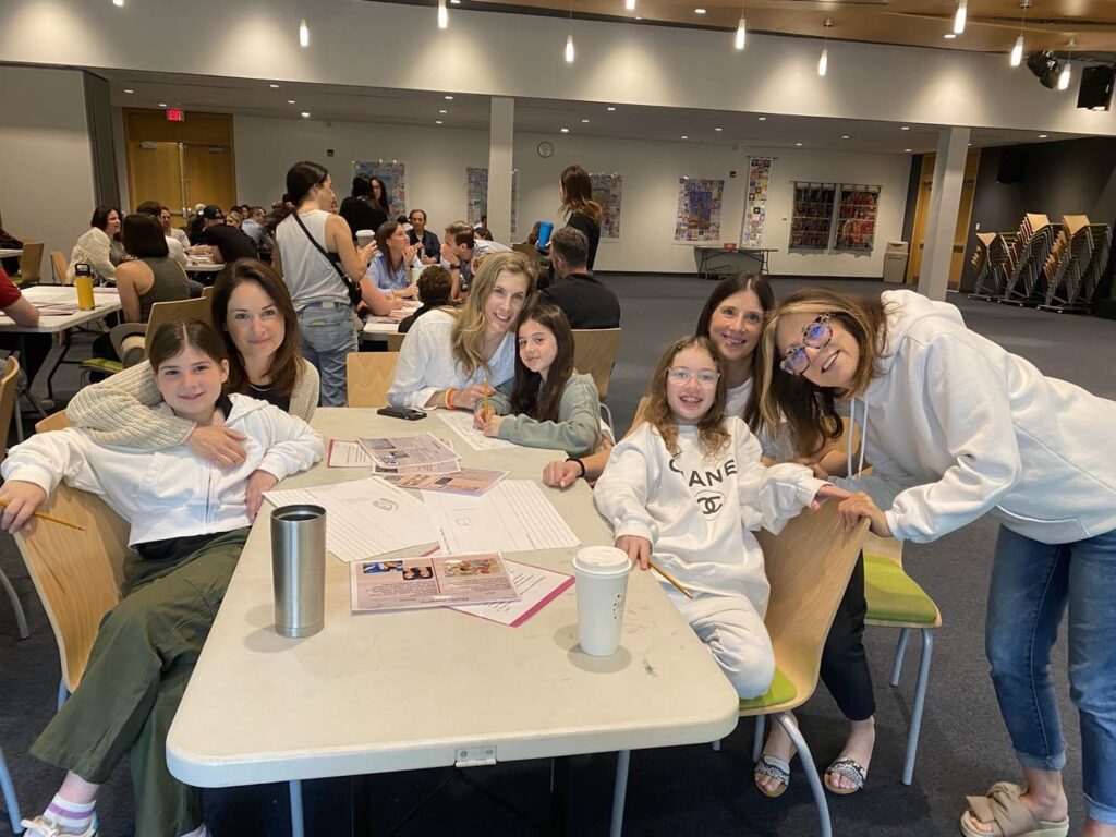A group of four mothers posing and embracing their children while sat around a table during an event in an auditorium.