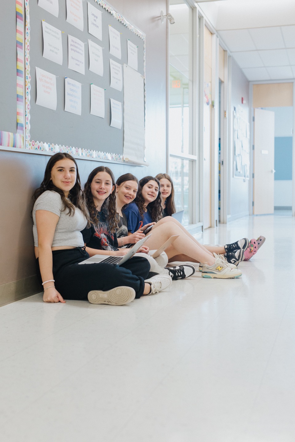 Five girls sitting against a wall in the school hall, working on a group project together.