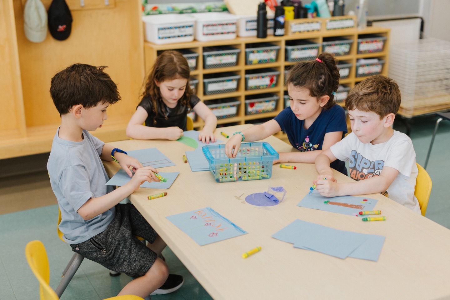 A group of 4 young students are sat at the same desk. They are sharing a basket of different coloured crayons and each drawing on a blue piece of paper.