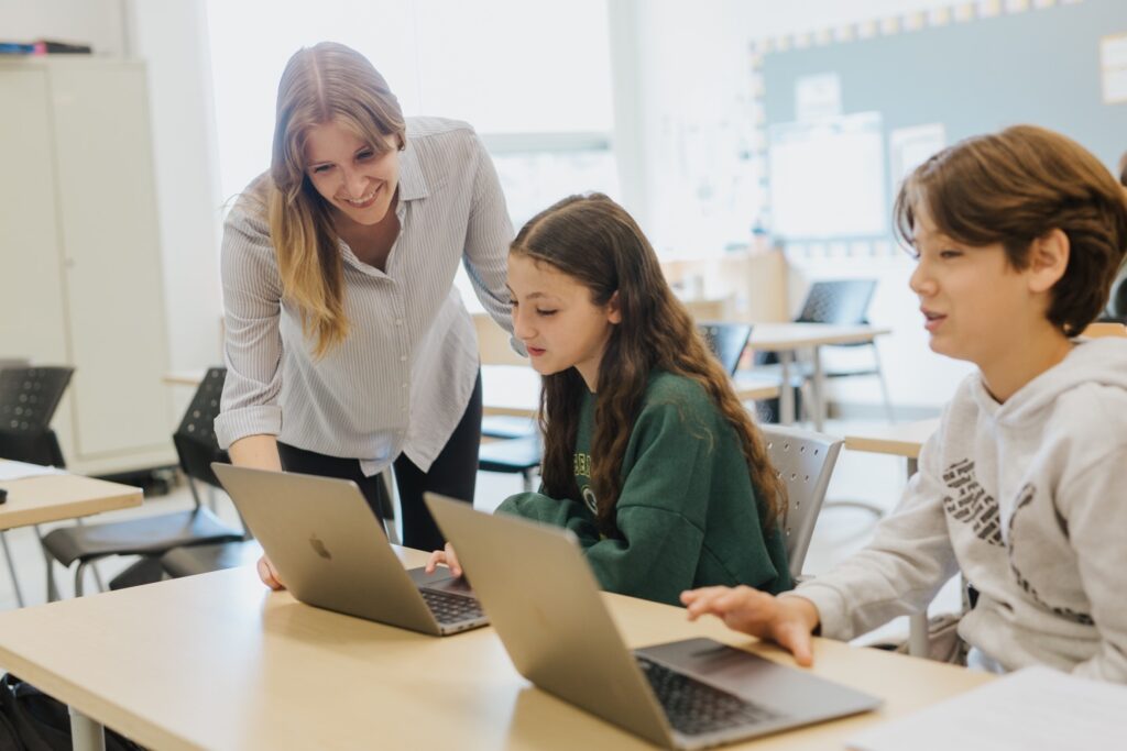 Two students are seated at a desk with Apple laptops. A teacher is giving friendly guidance to one of the students who is trying to solve a problem.