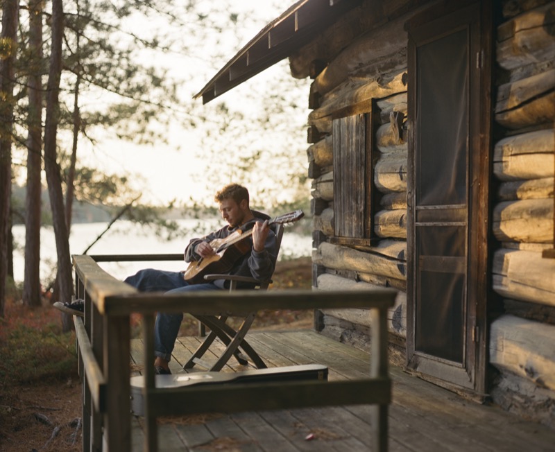 Nate Daviau sits on a deck of a wooden cabin next to a lake, He's an indie-folk singer/songwriter and a Leo Baeck alumni.