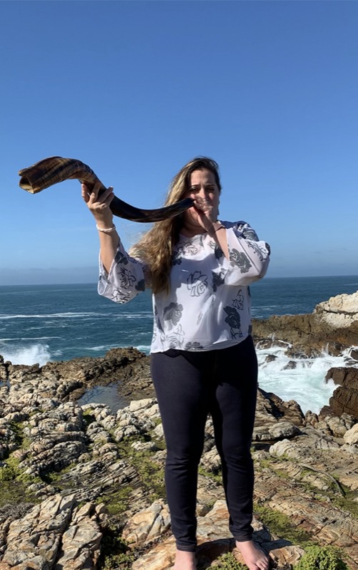 Emma Gottlieb blows a shofar next to a coastline. She is one of the first women rabbis at Temple Israel in Cape Town and a Leo Baeck alumni.