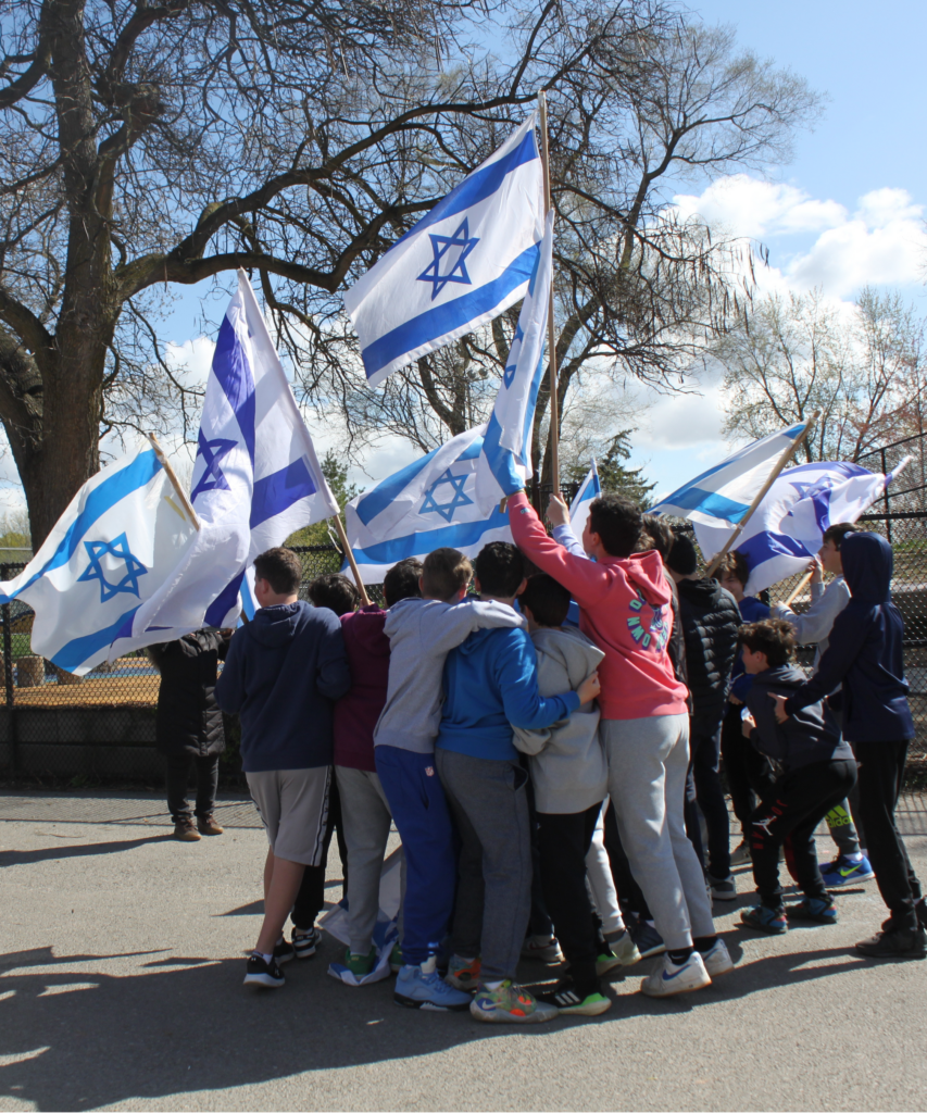 A large circle of Leo Baeck students gathered closely together holding several Israeli flags.