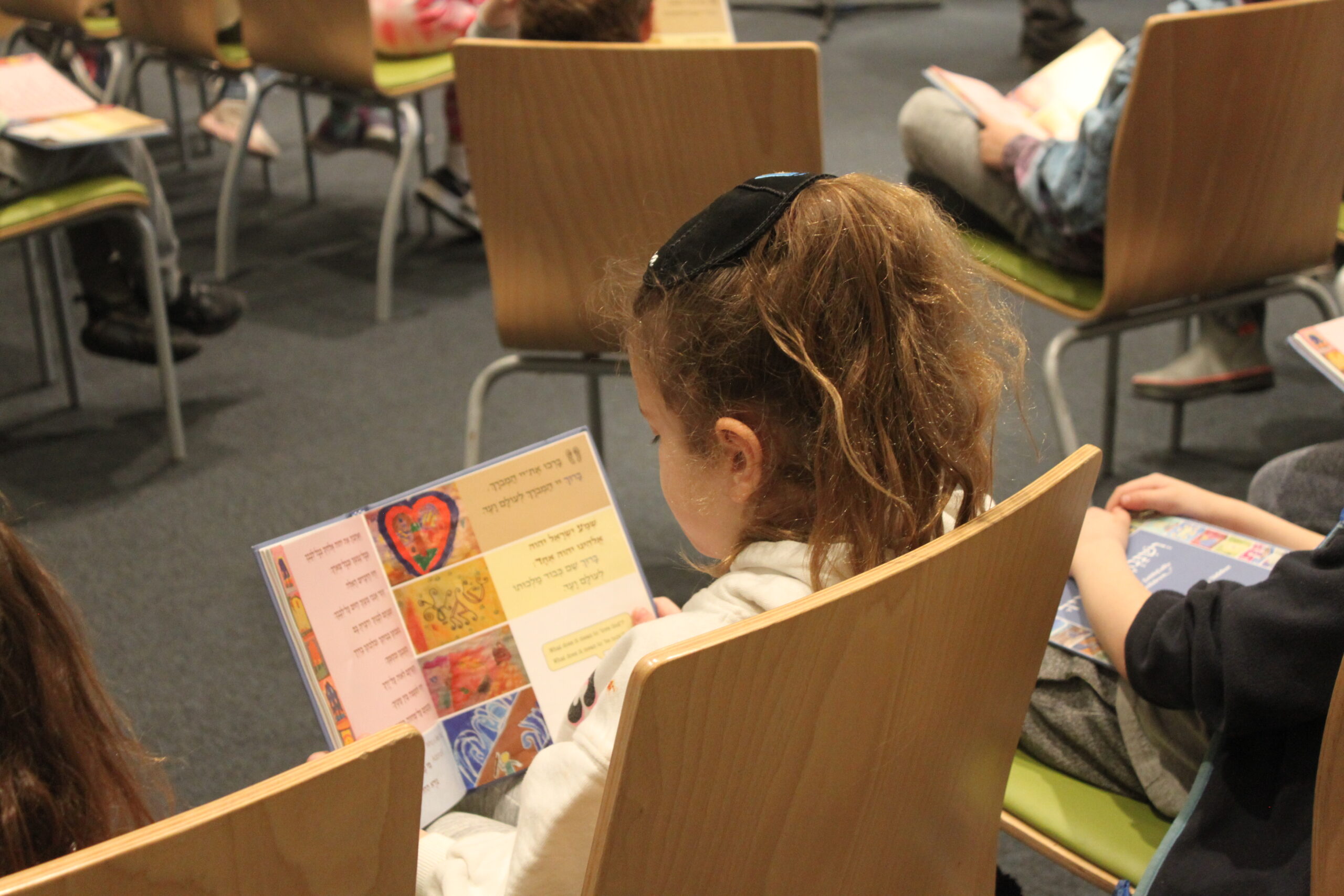 A young first or second grader sitting in a modern wooden chair, reading from a booklet along with many other children.