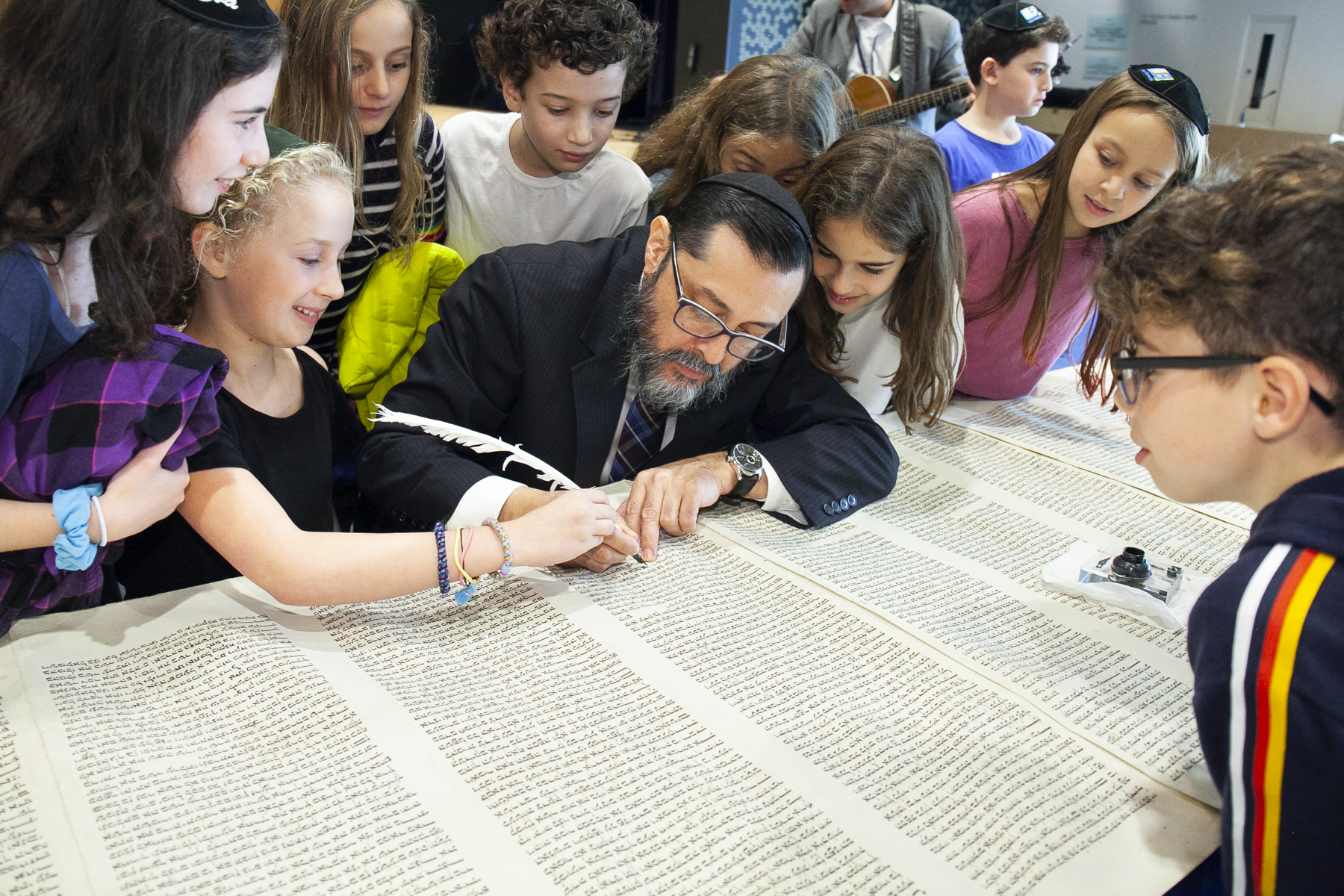 Rabbi Yochanan Salazar inscribing letters in a scroll together with children. Rabbit Salazar is surrounded by children watching the process with focus and interest.