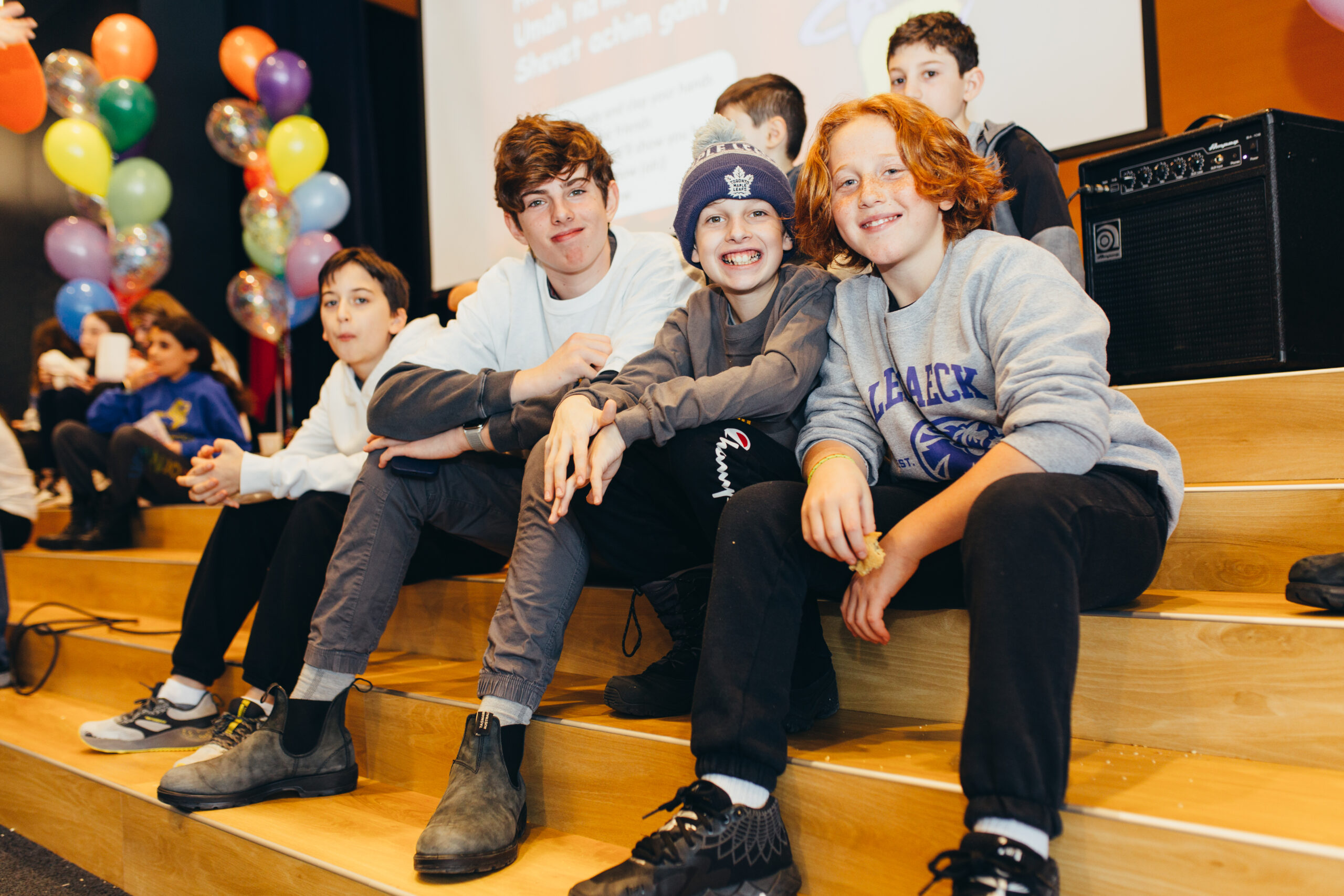 A group of students sitting on a stage at an event. There are balloons and a large screen in the background. The children are smiling naturally.